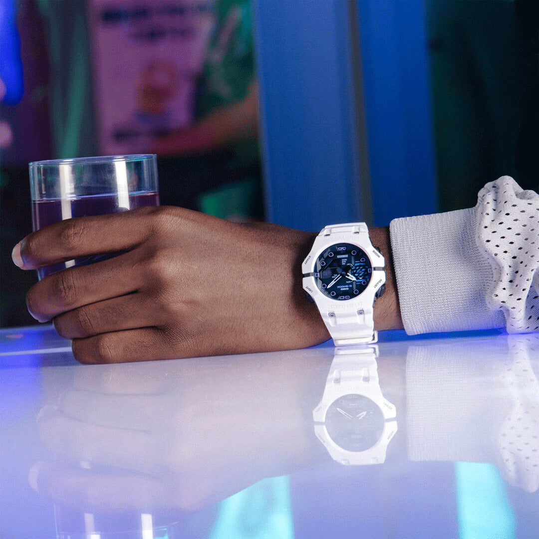 A white Casio watch with a blue dial worn on a person's wrist, resting on a reflective blue-lit table next to a glass of water.