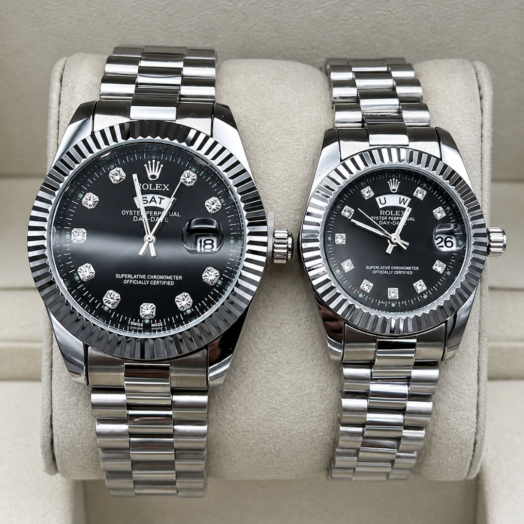 Two Rolex Datejust stainless steel watches with black dials and diamond hour markers, displayed side-by-side on a beige watch cushion. The watch on the left has a fluted bezel and a fitter-style bracelet, while the watch on the right has a rotating bezel and an Oyster bracelet.