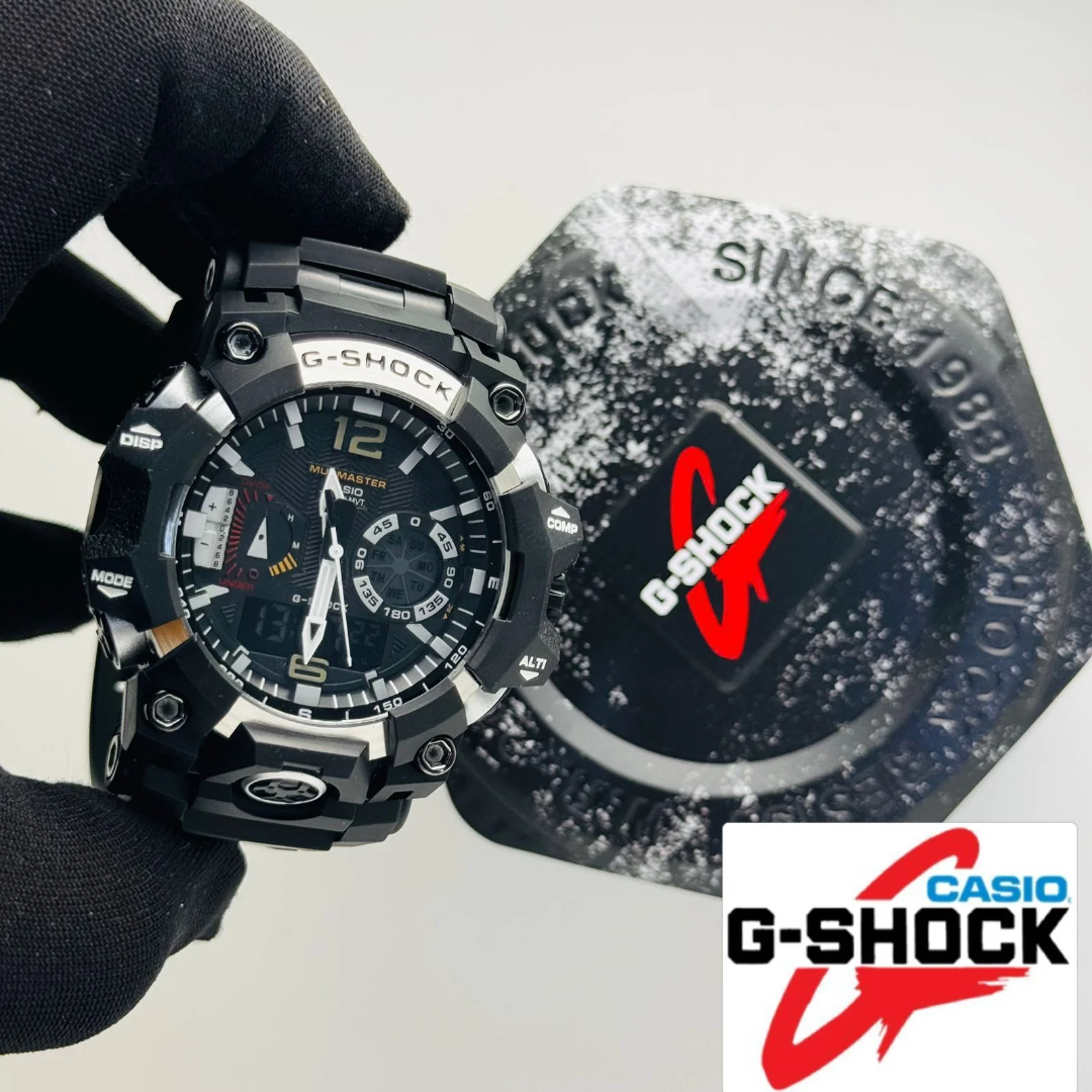 A black Casio G-Shock analog watch with a rugged design is held by a black gloved hand, with its matching black textured watch strap or accessory case placed behind it on a light gray background. A Casio G-Shock logo overlay is visible in the bottom right corner.