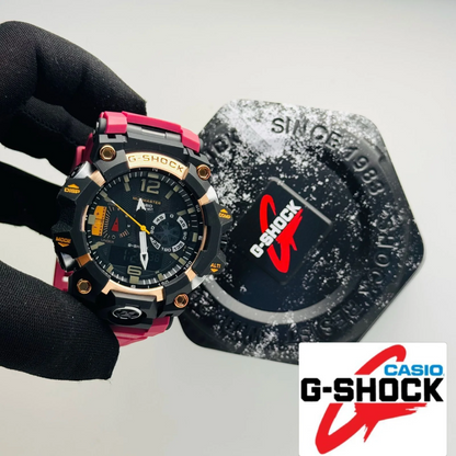 A Casio G-Shock watch with a black and pink strap and gold-tone accents, held by a gloved hand next to its branded hexagonal box on a light gray background.