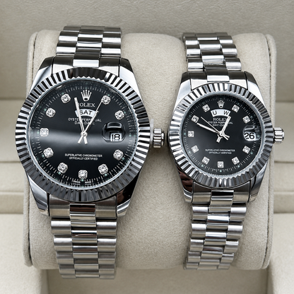 Two Rolex Datejust stainless steel watches with black dials and diamond hour markers, displayed side-by-side on a beige watch cushion. The watch on the left has a fluted bezel and a fitter-style bracelet, while the watch on the right has a rotating bezel and an Oyster bracelet.