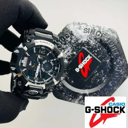 A black Casio G-Shock analog watch with a rugged design is held by a black gloved hand, with its matching black textured watch strap or accessory case placed behind it on a light gray background. A Casio G-Shock logo overlay is visible in the bottom right corner.